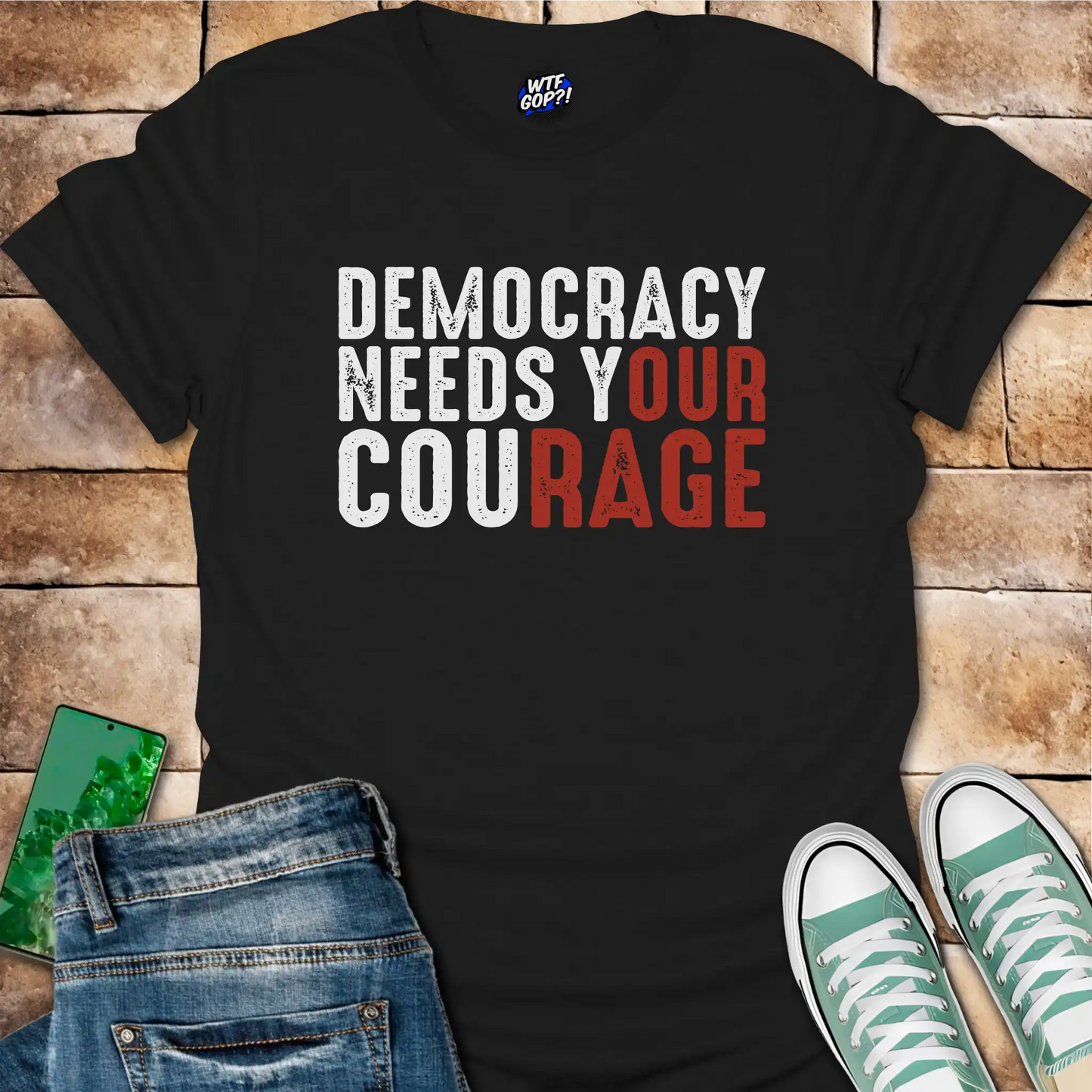 Black t-shirt with 'Democracy Needs Your Courage' text on a wooden surface with jeans and sneakers.