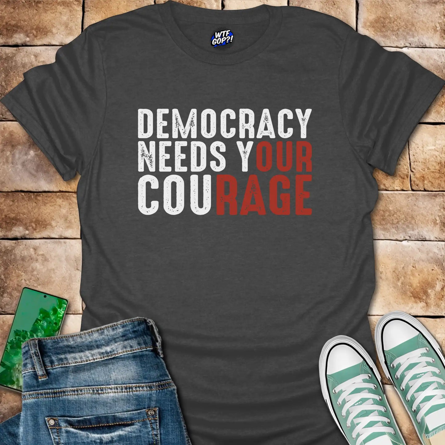 Dark gray t-shirt with 'Democracy Needs Your Courage' text on a wooden surface with jeans and sneakers.