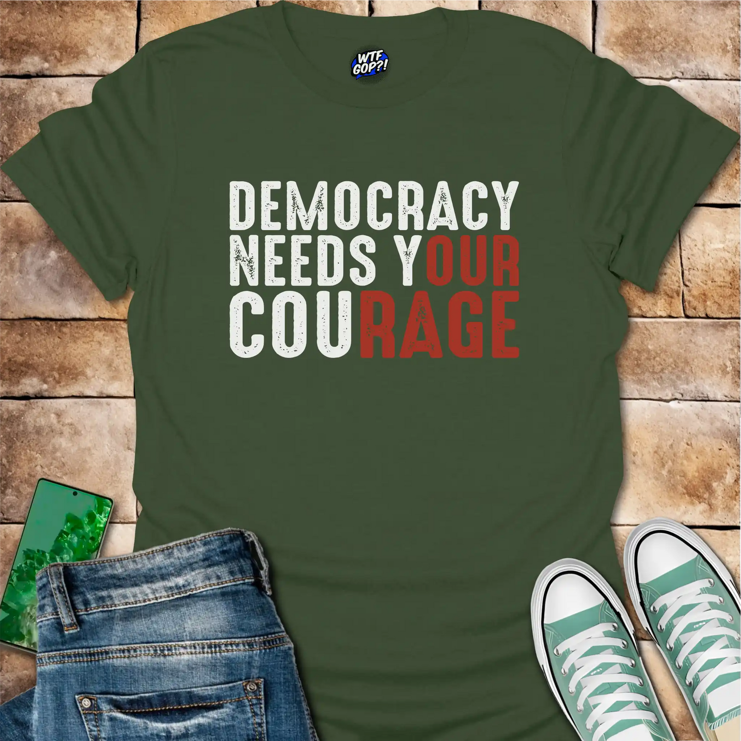 Green t-shirt with 'Democracy Needs Your Courage' text on a wooden surface with jeans and sneakers.